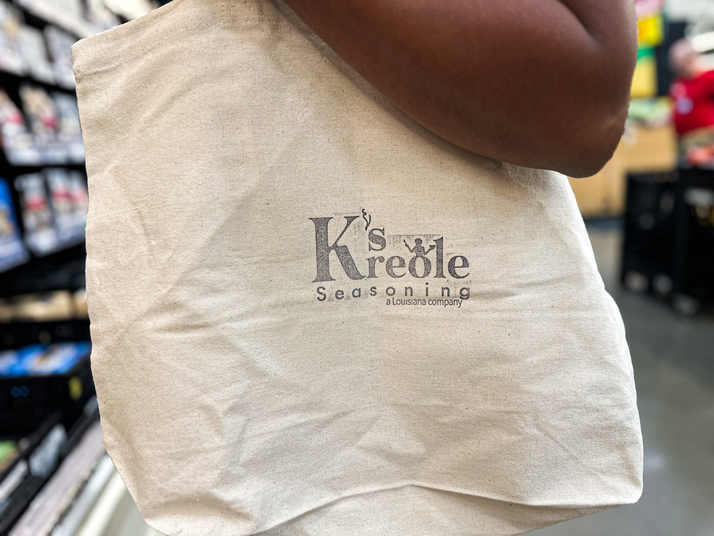 Beige reusable cotton grocery bag with 'K's Kreole Seasoning' logo held by a person in a store setting