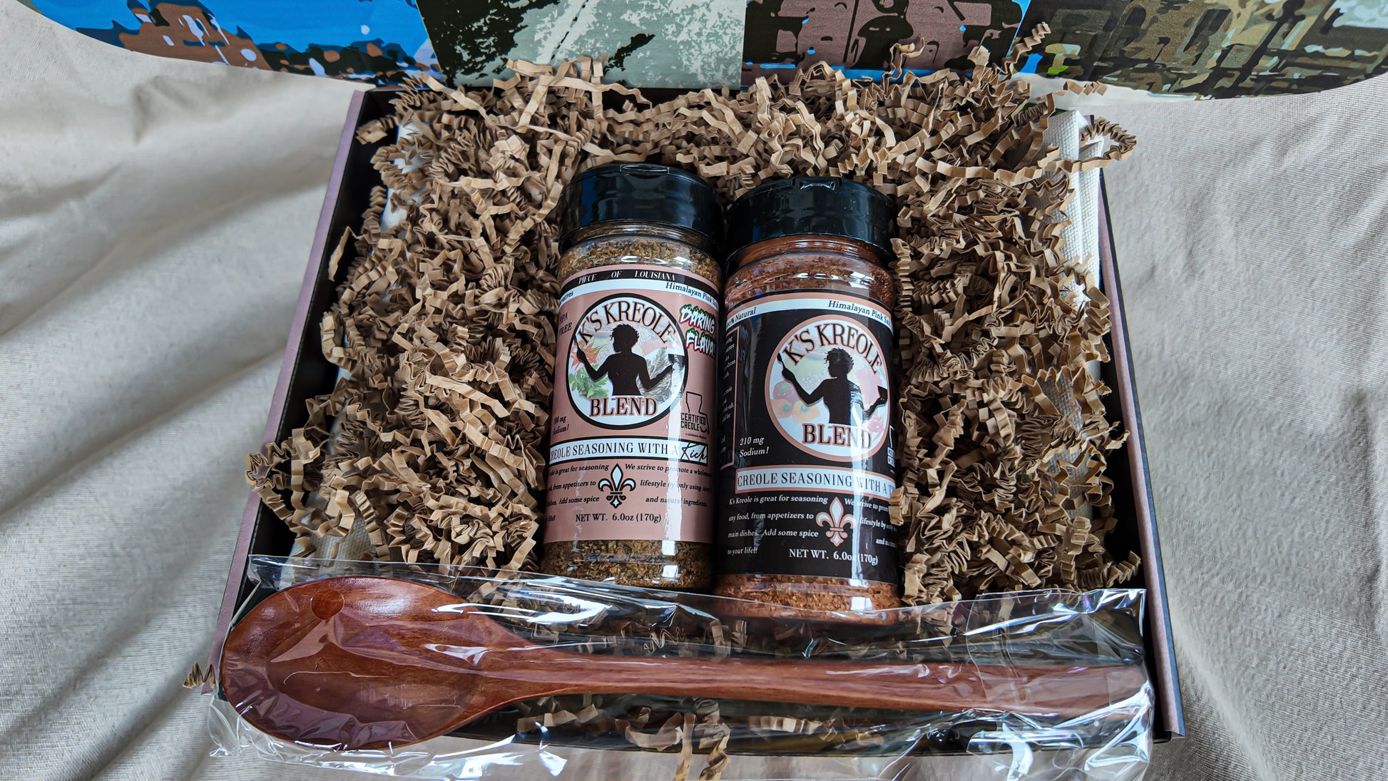Two bottles of K's Kreole Seasoning with a wooden spoon in a box with brown crinkle paper.